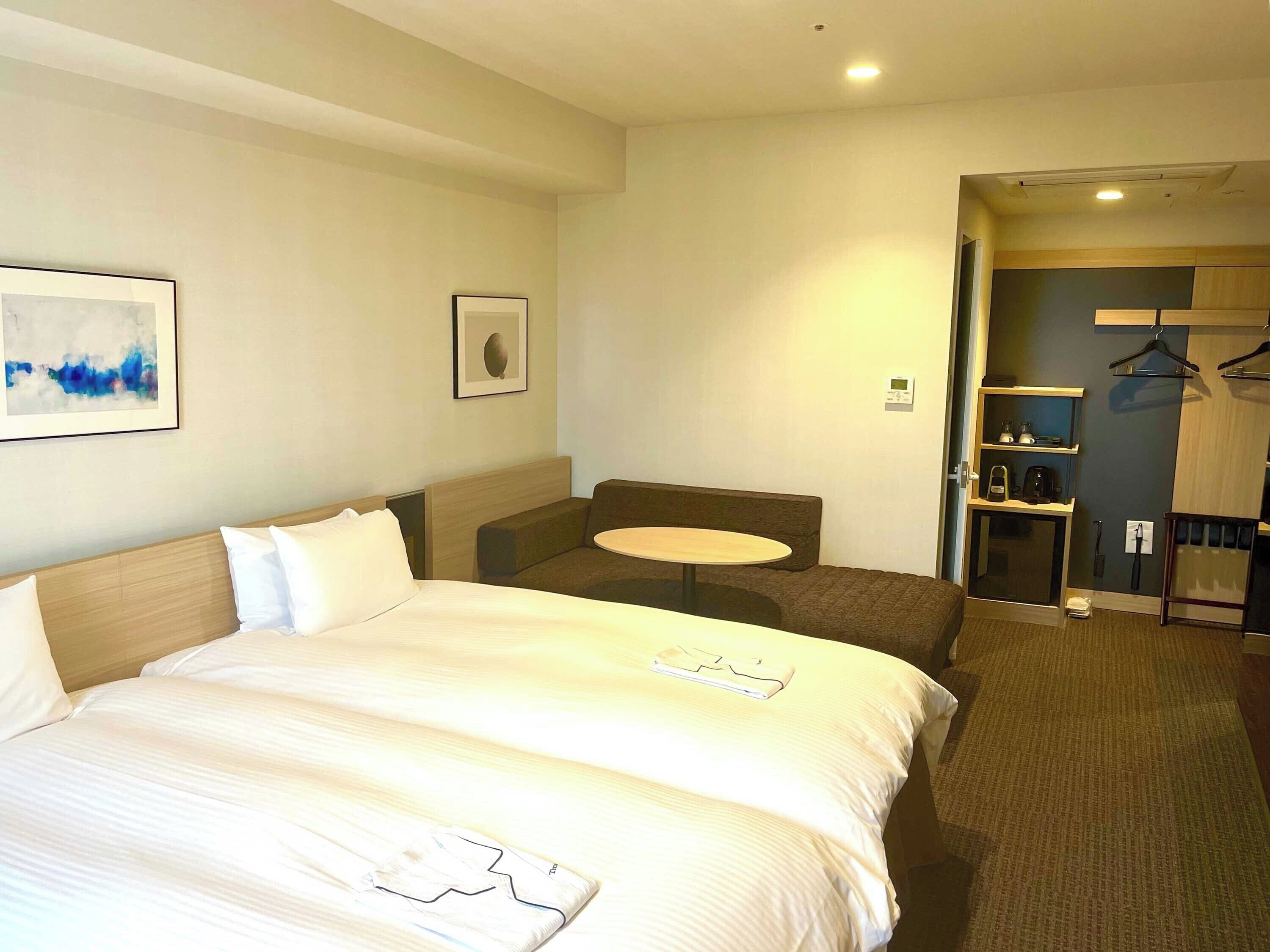 A hotel room with a large bed, a small round table, a sofa, and a wardrobe with hanging clothes.