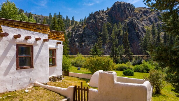 A rustic building with a white exterior and wooden roof, surrounded by a lush garden and a mountainous backdrop. - Adobe House on Taylor River | 3 Bedroom | River Access | Gas Grill | Yard (Almont)