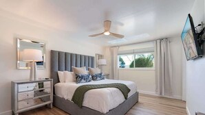 A modern bedroom with a large bed, a ceiling fan, a bedside table with lamps, and a window with curtains.