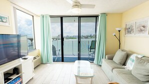 A living room with a television, a glass-top coffee table, a sofa, and a view of a cityscape through a sliding glass door. - # 902 Gulf View at Lovers Key Beach Club (Bonita Springs)