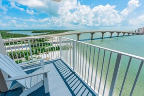 A balcony with a view of a bridge over water, a chair, and a pathway leading to a beach.