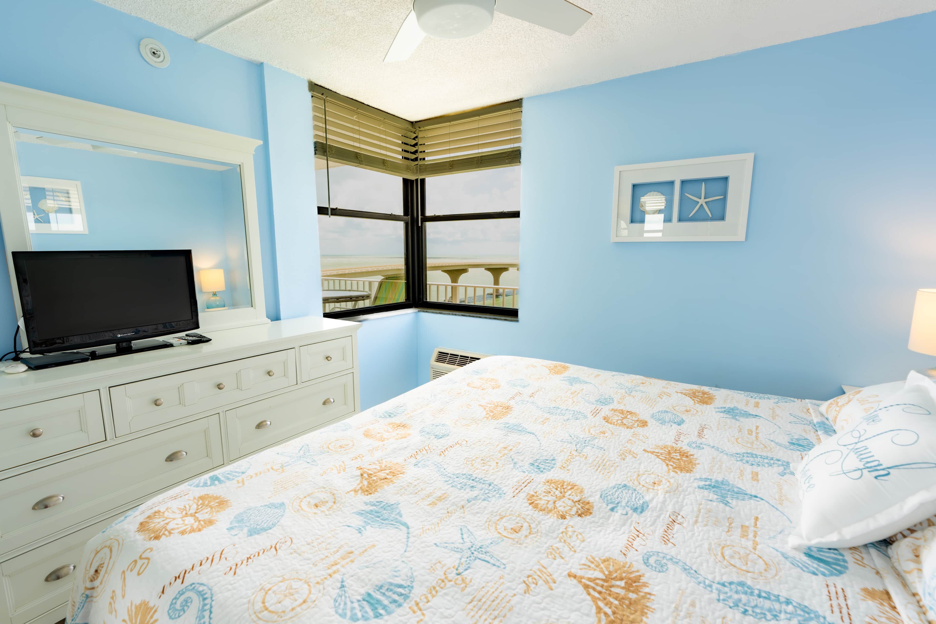 Une chambre à coucher avec un lit, un téléviseur posé sur une commode, une fenêtre avec des stores et un mur orné de tableaux.