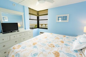 A bedroom with a bed, a TV on a dresser, a window with blinds, and a wall with framed pictures.