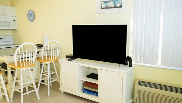 A room with a white cabinet, a flat-screen TV, two white chairs, a microwave, a clock, and a framed picture on the wall. - # 902 Gulf View at Lovers Key Beach Club (Bonita Springs)