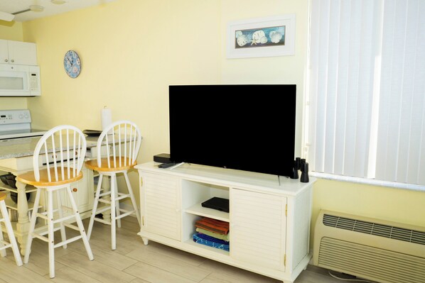 A room with a white cabinet, a flat-screen TV, two white chairs, a microwave, a clock, and a framed picture on the wall.