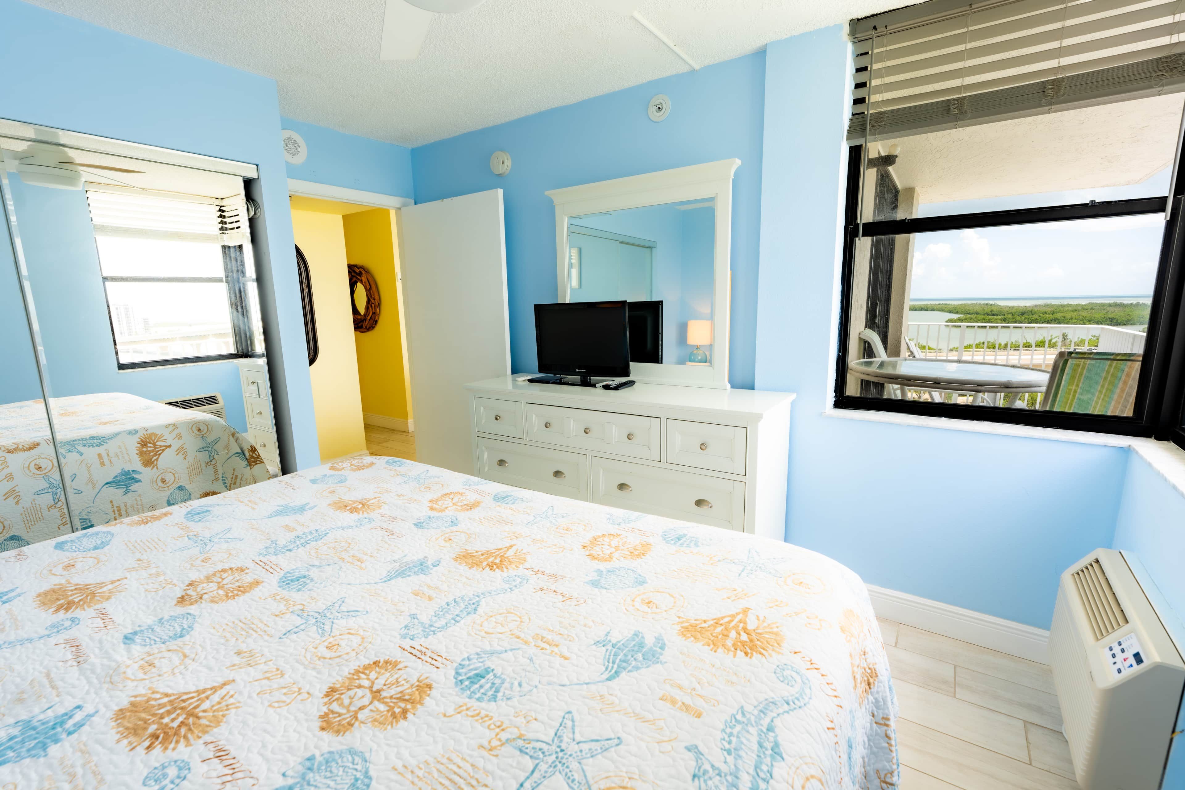 A hotel room with a bed, a television, a dresser, and a window with a view of the ocean.