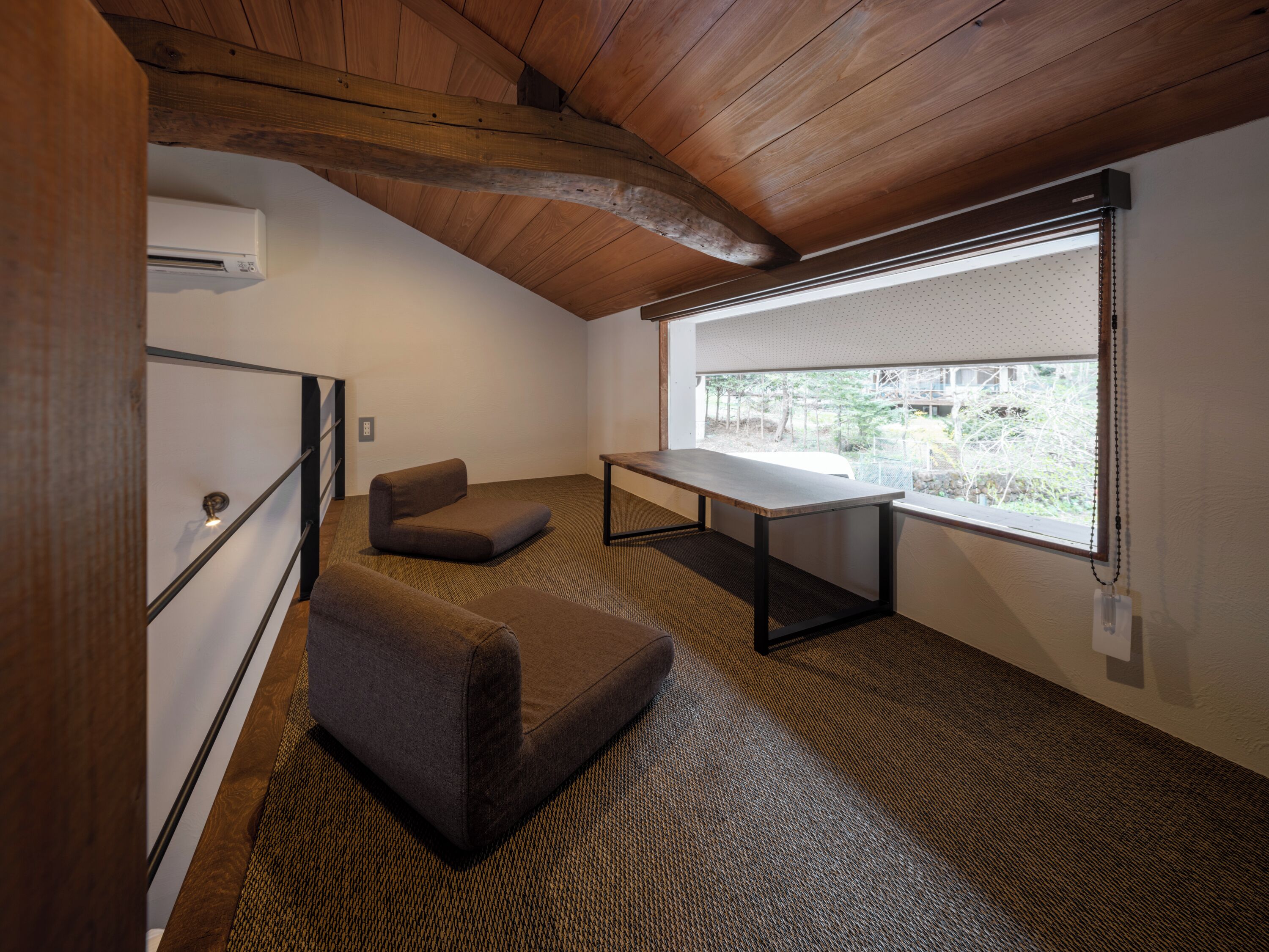 A room with a sloped ceiling, a large window with blinds, a sofa, a small table, and a wall-mounted air conditioner.
