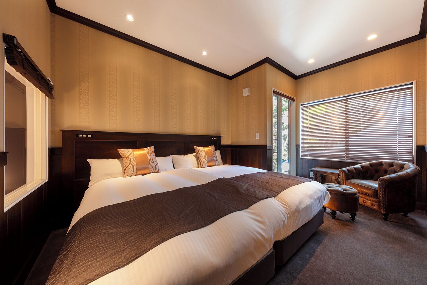 A bedroom with a large bed, a wooden headboard, a window with blinds, and a leather armchair.