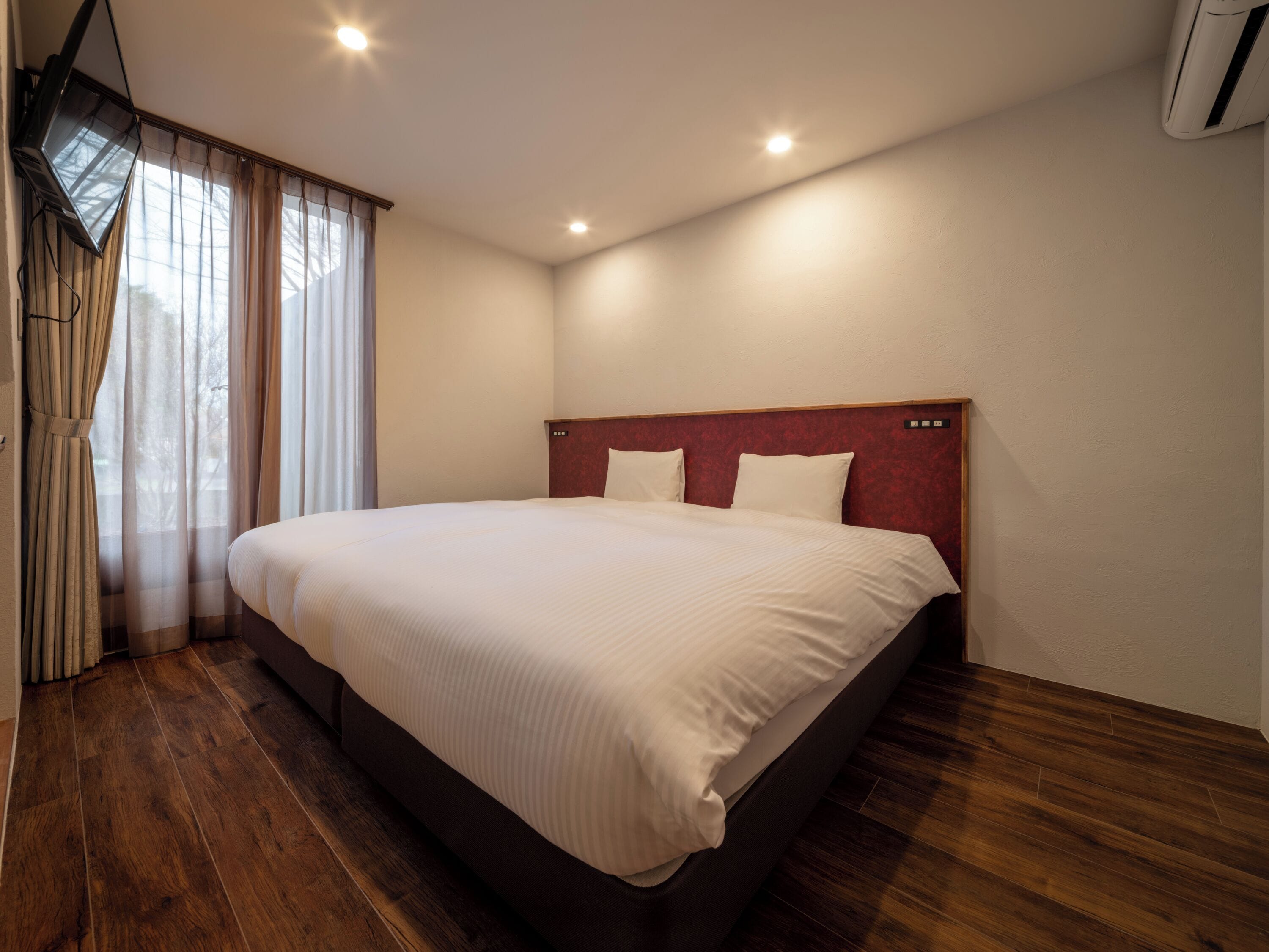 Ein Hotelzimmer mit einem großen Bett, Holzboden, einem roten Kopfteil und einem an der Wand montierten Fernseher.