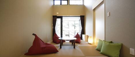 A room with tatami mat flooring, a low table, red and green cushions, and a view through a large window.