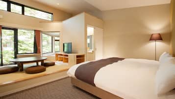 A modern hotel room with a large bed, a television, a desk, and a view of greenery outside.