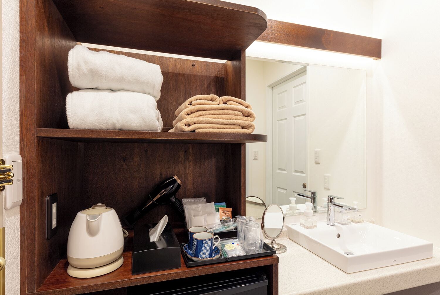Un baño de hotel con un estante de madera que contiene toallas, una cafetera, un secador de pelo y artículos de aseo.