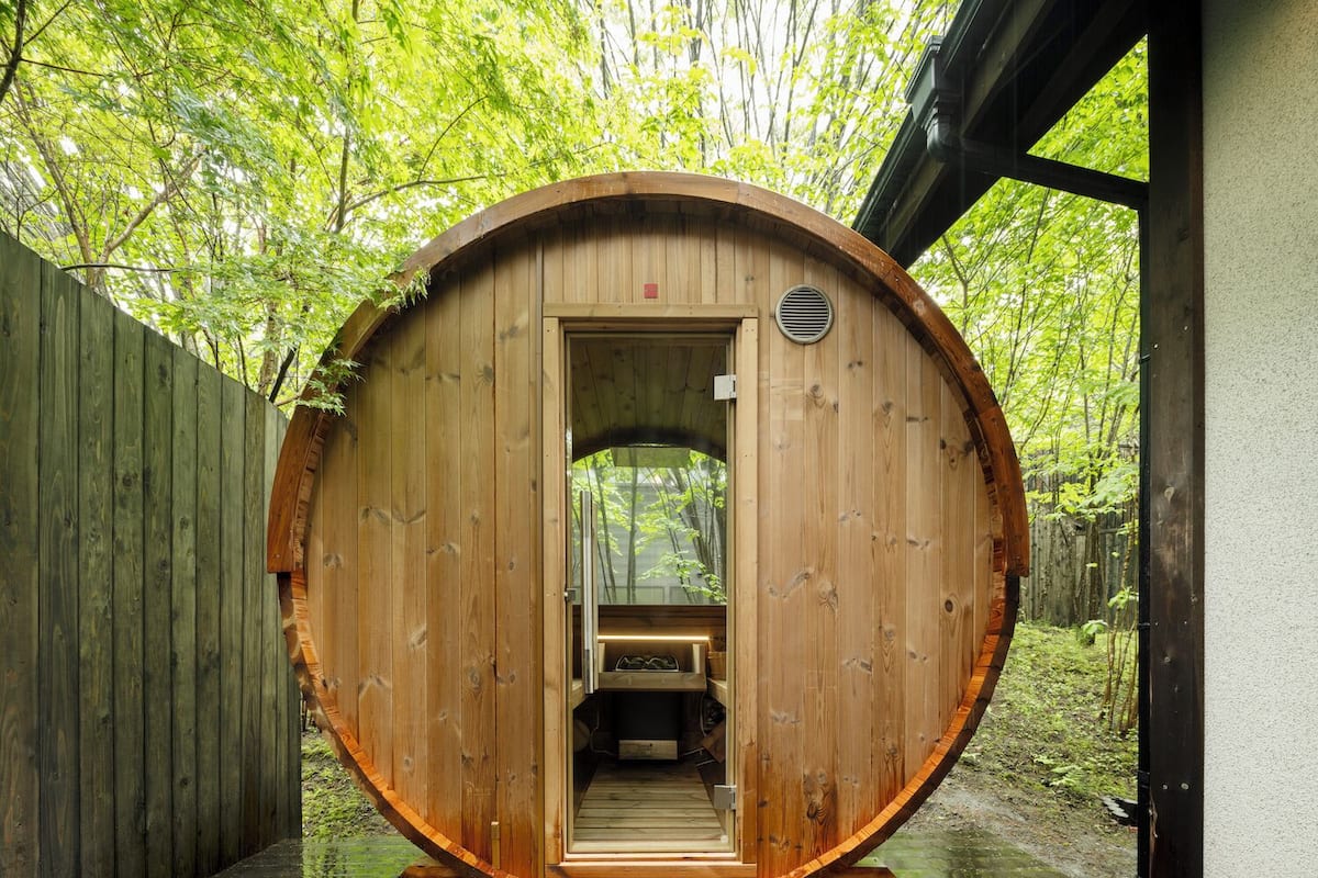 Eine Holzsauna mit einer Sitzbank im Inneren, umgeben von einem Holzzaun und Bäumen.