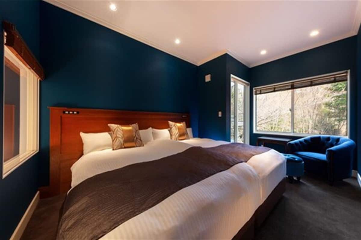 Ein großes Bett mit hölzernem Kopfteil, ein Nachttisch und ein Stuhl in einem Zimmer mit dunkelblauen Wänden und einem großen Fenster.
