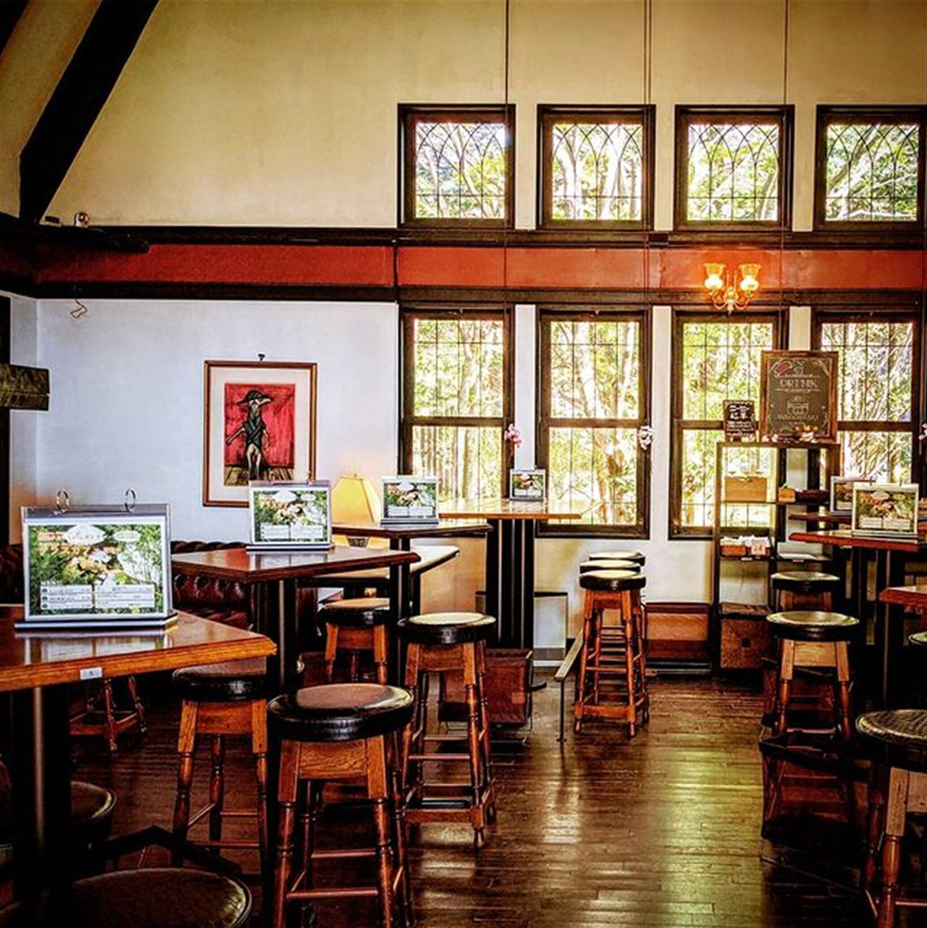 Un restaurante con mesas y sillas de madera, grandes ventanales con follaje en el exterior y obras de arte enmarcadas en las paredes.