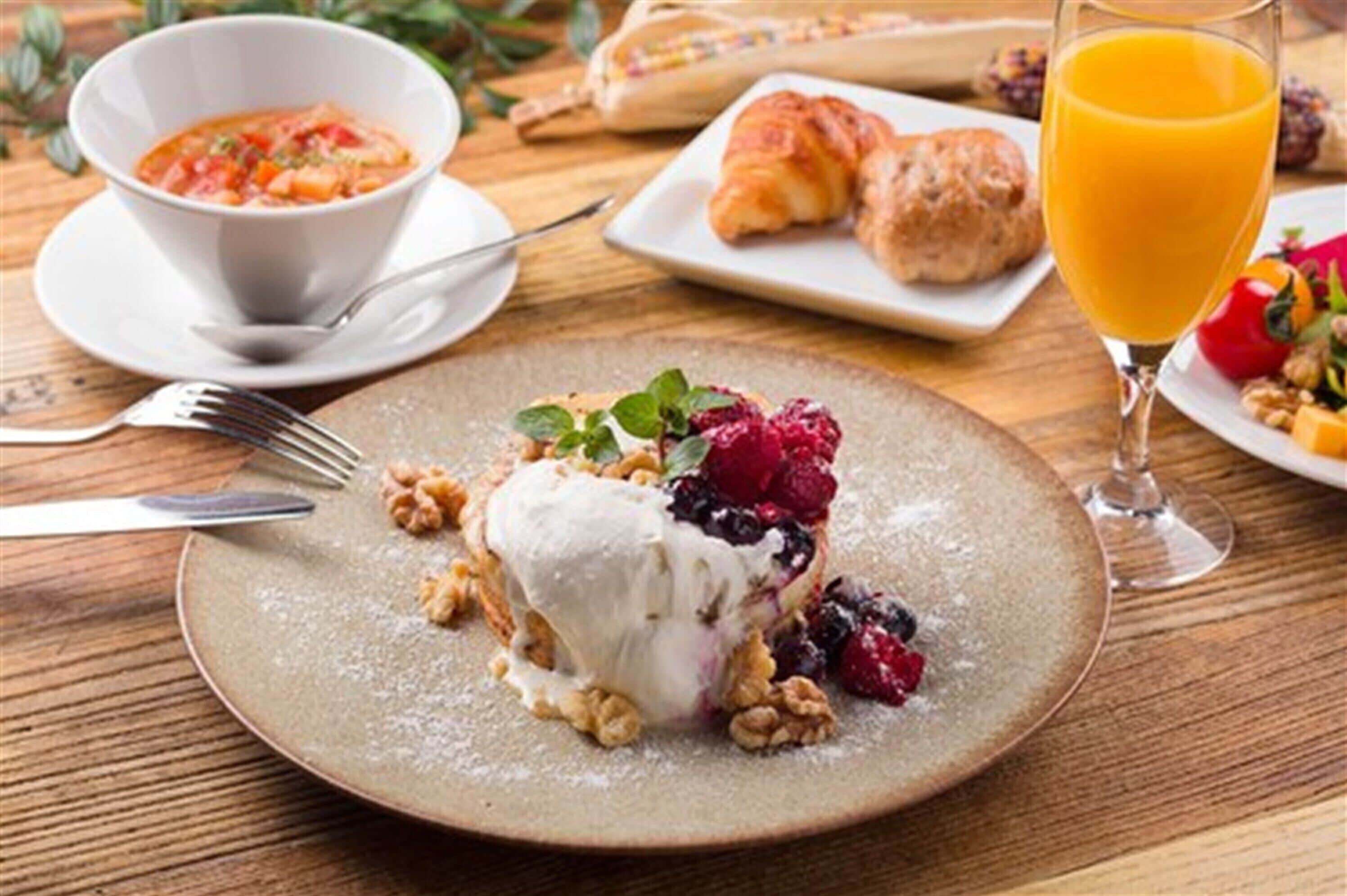 A breakfast setting with a plate of dessert, a bowl of soup, croissants, and a glass of orange juice.