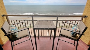 Ein Balkon mit zwei Stühlen und einem Tisch, der Blick auf einen Strand mit Wellen und einem klaren Himmel bietet.
