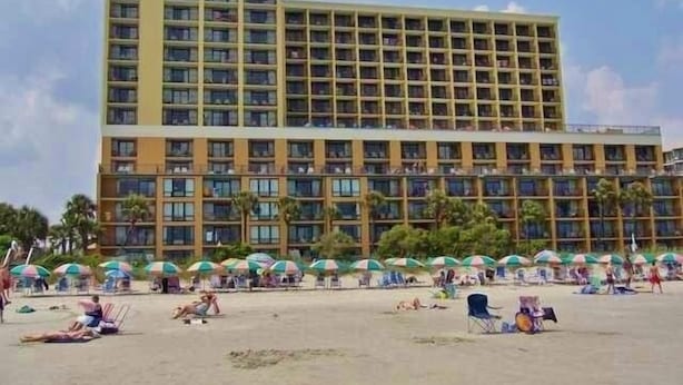 Ein Hotel mit mehreren Etagen, einem Strand davor und Sonnenschirmen.