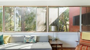 A cozy living room with a grey sofa, a small wooden table, and a lamp. Large windows offer a view of trees and a building outside. - Silver Lake Mid-century Nestled in the Treetops (Los Angeles)