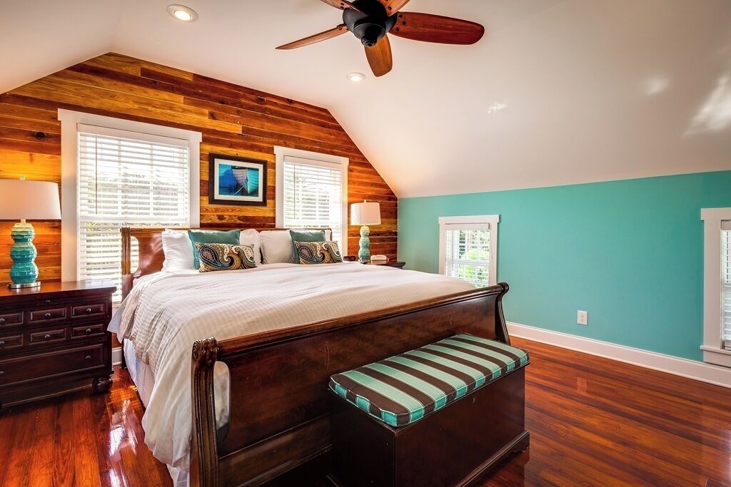 A bedroom with a wooden ceiling, a large bed, two windows with blinds, and a ceiling fan.