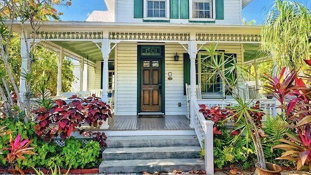 Una casa a due piani con un portico, una porta d'ingresso con la targa '1501', e un giardino curato con diverse piante.
