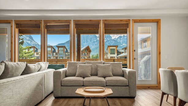 Un salon moderne doté d’un grand canapé d’angle, d’une table basse en bois et offrant une vue sur les maisons de montagne grâce à de grandes fenêtres.