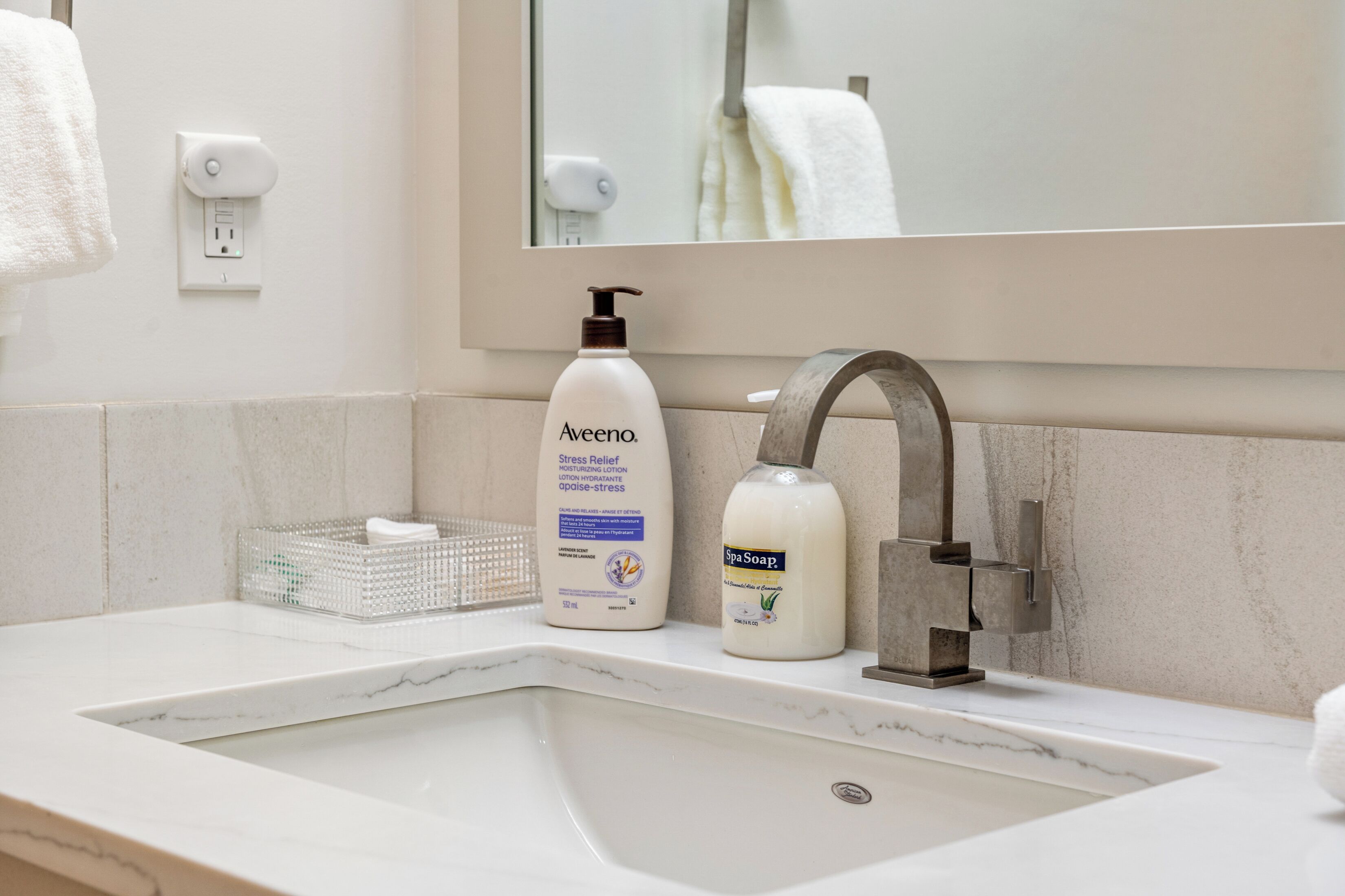 Ein Badezimmerwaschbecken mit Marmorplatte, ein Spiegel, ein Handtuchhalter und Pflegeprodukte, darunter Aveeno Lotion und Spa-Seife.