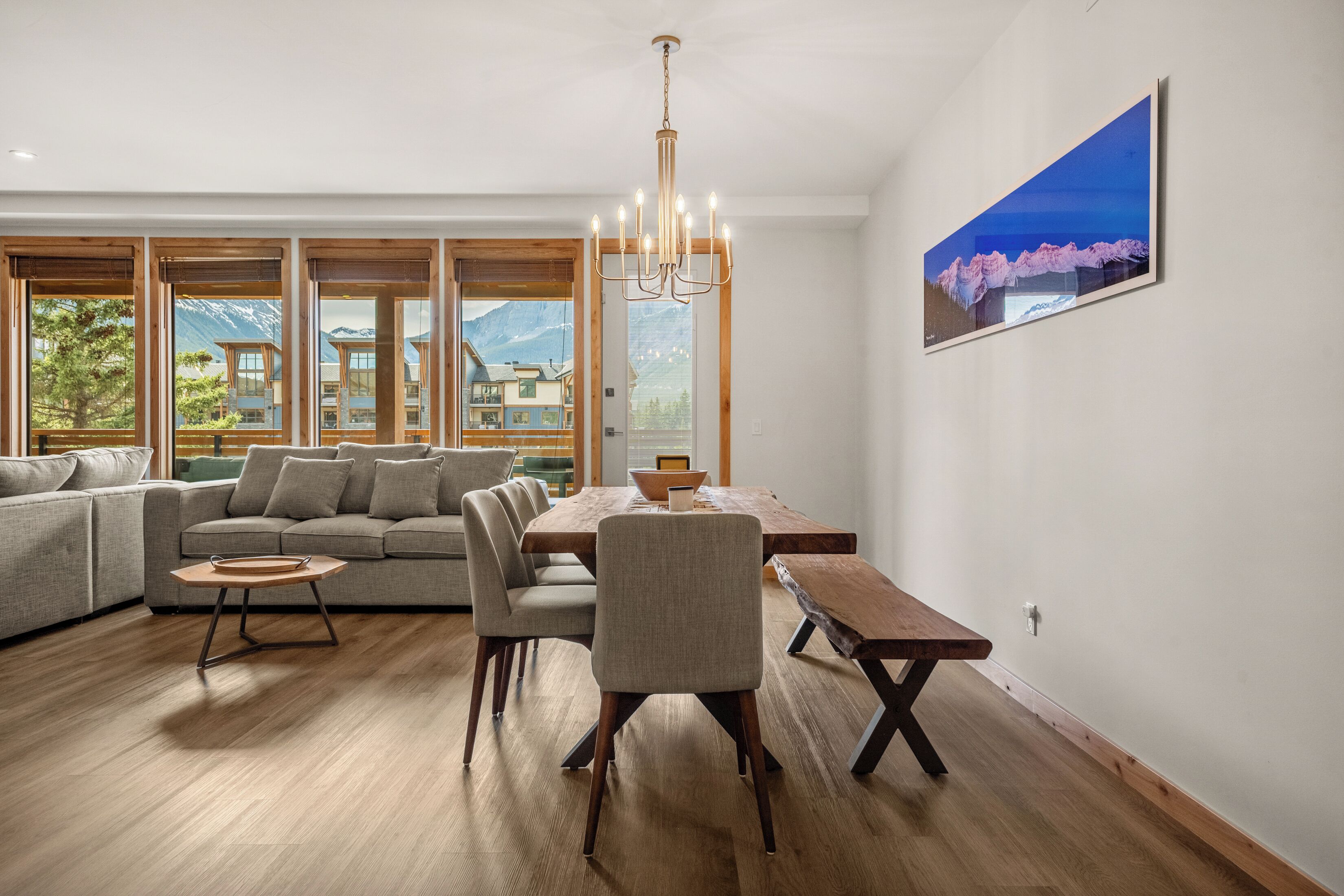 A modern living room with a wooden dining table, a sofa, and a large window with a mountain view.