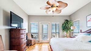 A bedroom with a TV, wooden dresser, a bed with white bedding, a ceiling fan, and a view through the door.