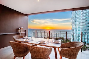 A balcony with a table set for two, overlooking a sunset over the sea, with a high-rise building in the background.