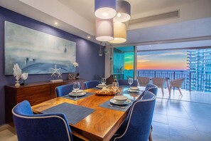 A modern dining area with a wooden table, blue chairs, and a view of the sunset through large windows.