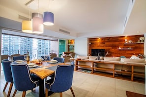 A modern living room with a dining area, large windows, and a wooden accent wall. - Luxurious, Beachfront 2bed/2 bath Condo with Designer Finishes (Puerto Vallarta)