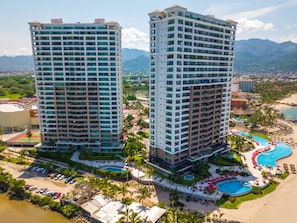Two high-rise buildings with multiple balconies, a swimming pool area with umbrellas, and a beach with parked cars.