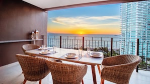 Un balcon avec une table dressée pour le dîner, des chaises en osier et une vue sur le coucher de soleil sur l’océan.