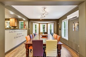 A dining room with a large wooden table, multiple chairs, a chandelier, and a kitchen area with cabinets and a window.