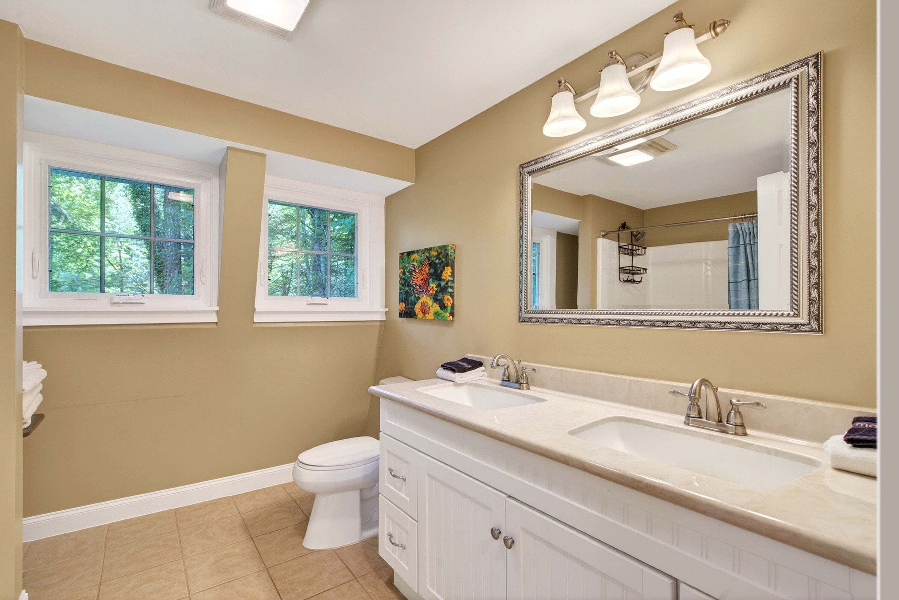 Une salle de bain avec un meuble-lavabo blanc, un grand miroir et deux fenêtres.