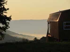A wooden cabin with a sloped roof, situated in a rural setting with a view of mist-covered hills in the background. - Lost Valley View Cabin (Ponca)