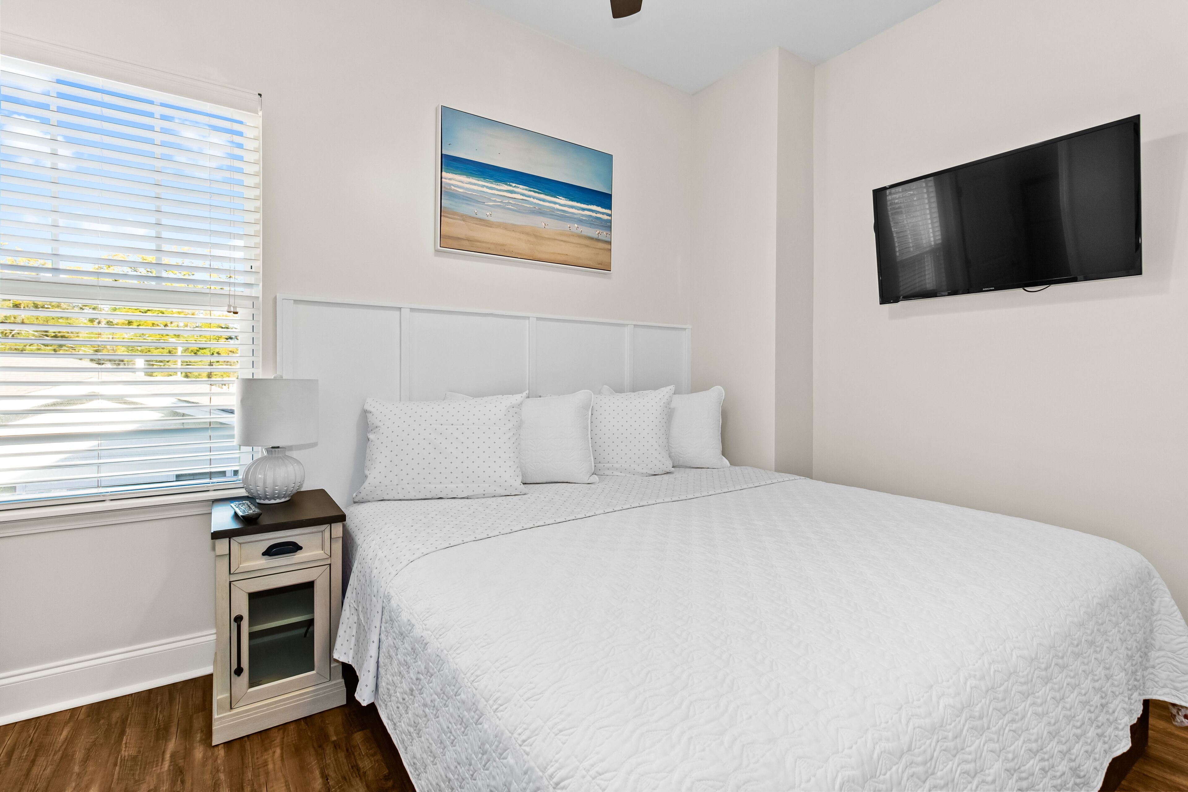 A bedroom with a large bed, a nightstand with a lamp, a TV mounted on the wall, and a beach painting above the bed.