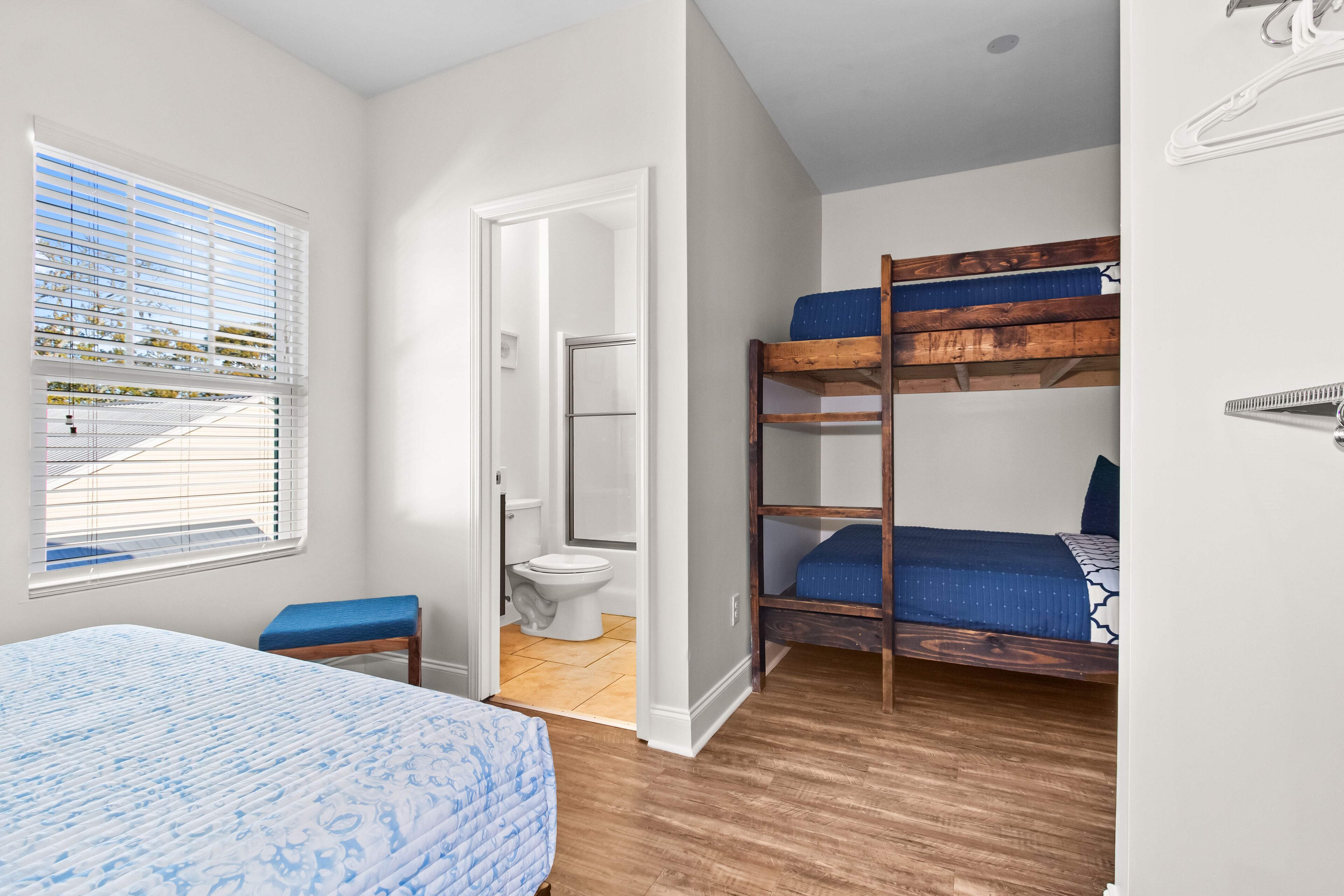 A compact bedroom with a bunk bed, a single bed, a bathroom with a toilet and shower, and a window with blinds.