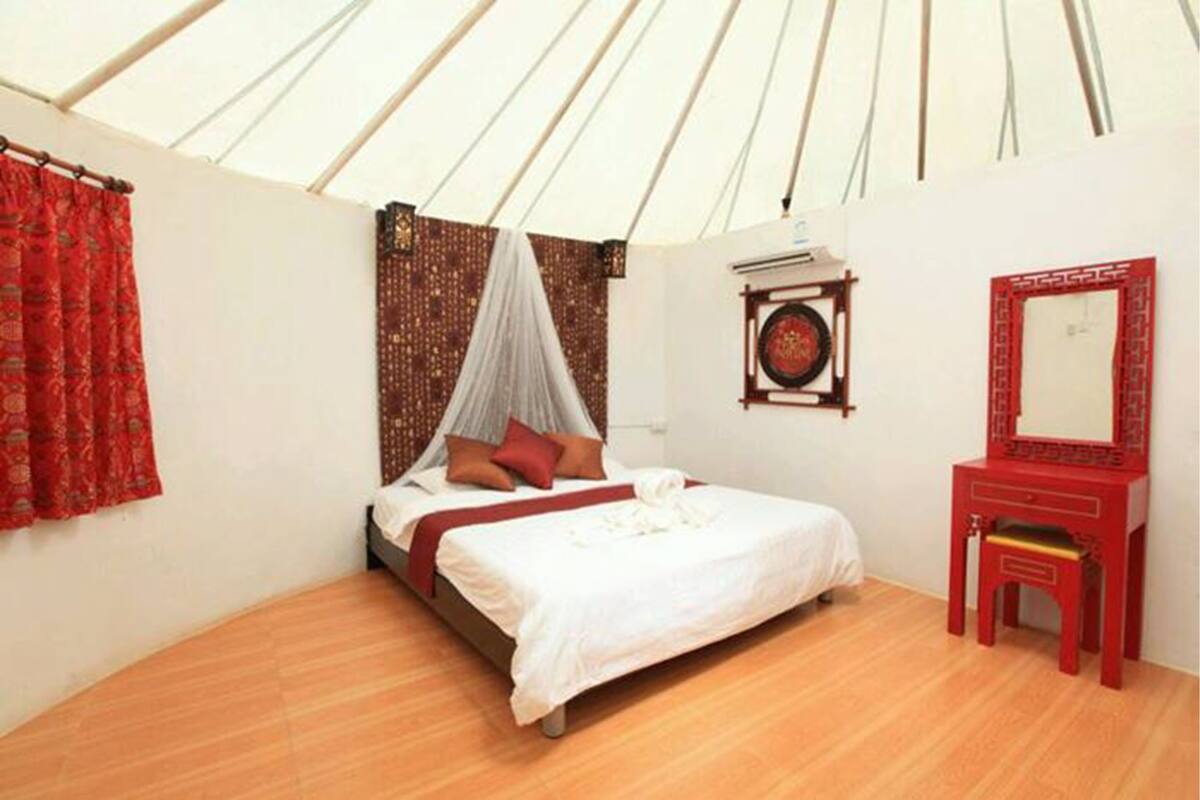 Un lit simple avec un baldaquin, une chaise rouge, un miroir et une étagère fixée au mur, sur laquelle est posé un objet décoratif.