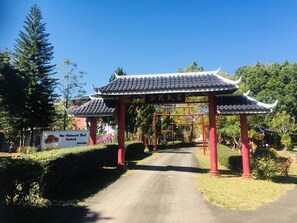 Une porte chinoise traditionnelle, avec un toit recouvert de tuiles et des piliers rouges, qui mène à un espace vert agrémenté de haies et d’arbres bien entretenus.