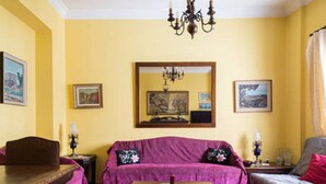 Una habitación con una pared amarilla, una mesa de madera, un sofá morado y una araña.