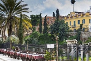 A row of outdoor tables with a fence in the foreground, a palm tree, and buildings in the background. - The best shelter in the historical, commercial and nightlife center of the city! (Athina)