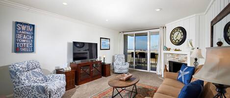 A living room with a sofa, armchair, coffee table, and a television. There is a fireplace and a large window with a view.