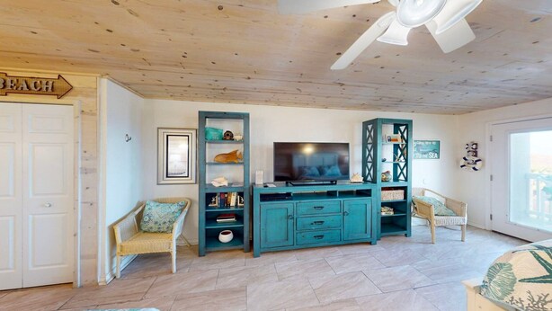 A living room with a television, teal cabinets, wicker chairs, and a ceiling fan. - Studio Condo in Town, Near Beach, Shared Pool and Hot Tub (Port Aransas)