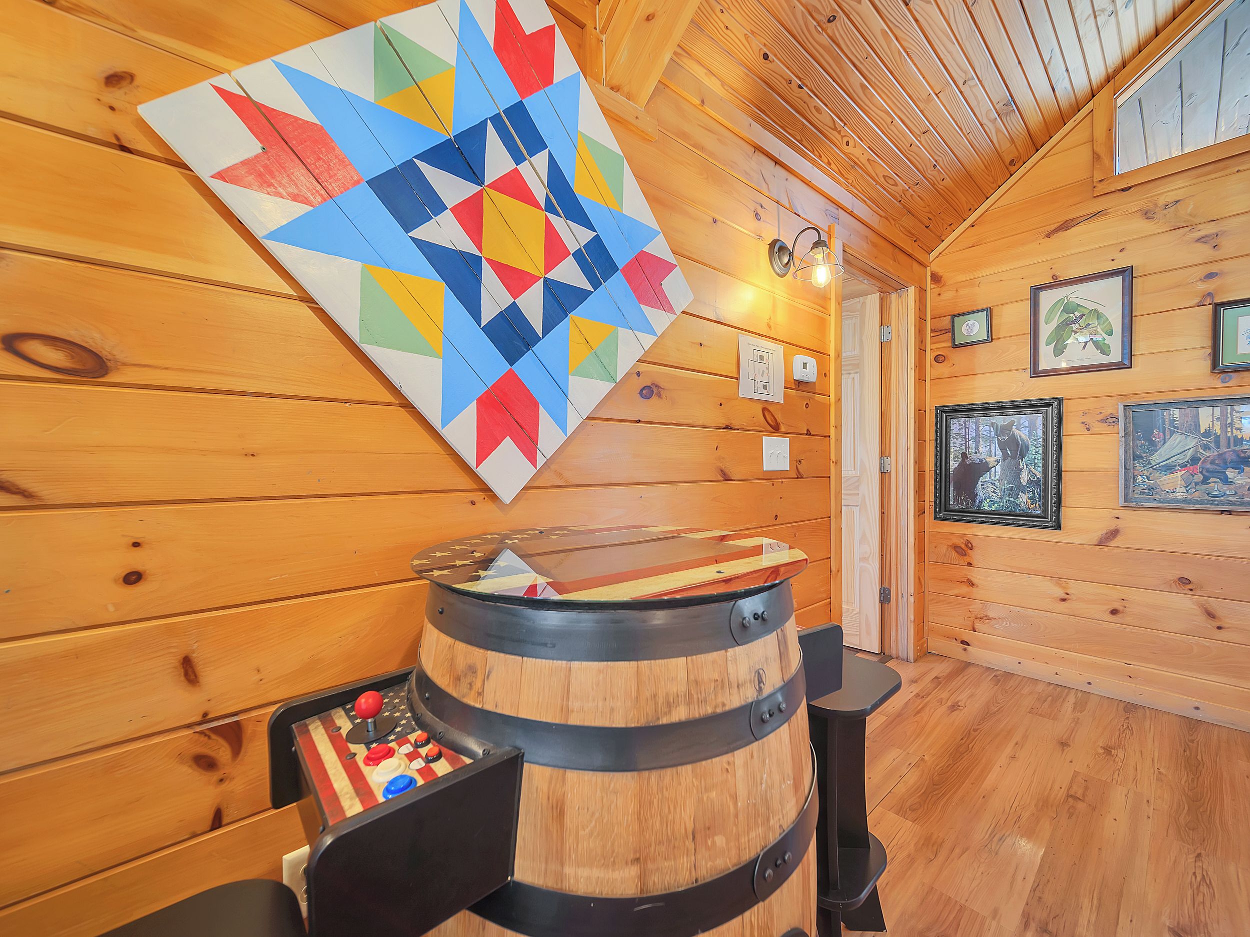 A wooden interior with a quilt hanging on the wall, a barrel with a table, and framed pictures on the wall.