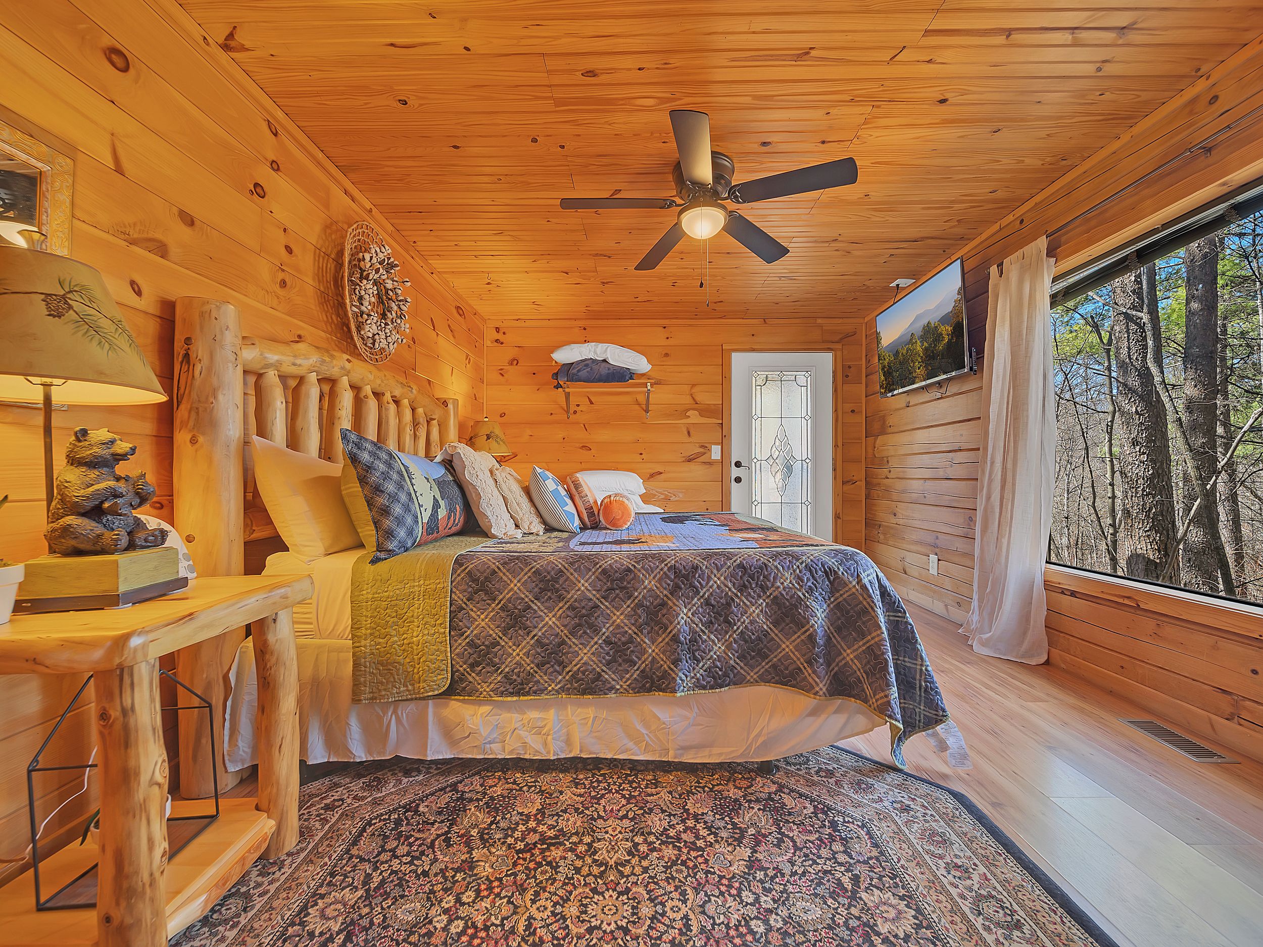 A cozy cabin room with a bed, a ceiling fan, a wooden headboard, a rug, and a window with a view of trees.