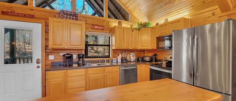 A wooden cabin kitchen with a large wooden table, stainless steel appliances, and a view of the forest through the window.