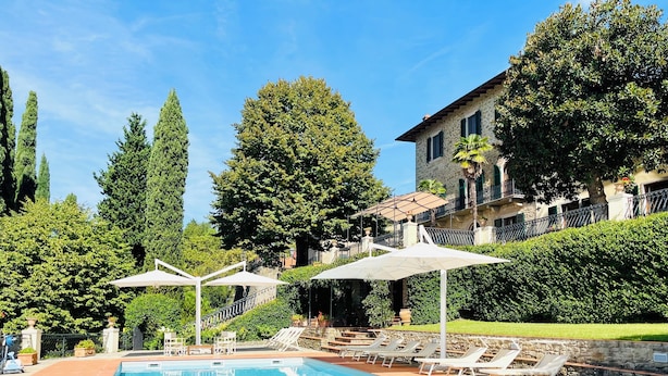 A swimming pool with lounge chairs and umbrellas, surrounded by greenery and a stone building.
