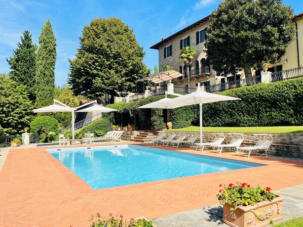 A swimming pool with lounge chairs and umbrellas, surrounded by greenery and a stone building. - A Panoramic Tuscan Luxury - Villa Maria in Fronzano (Reggello)